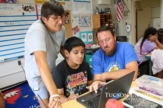 Teachers stand behind a student helping to use the computer.