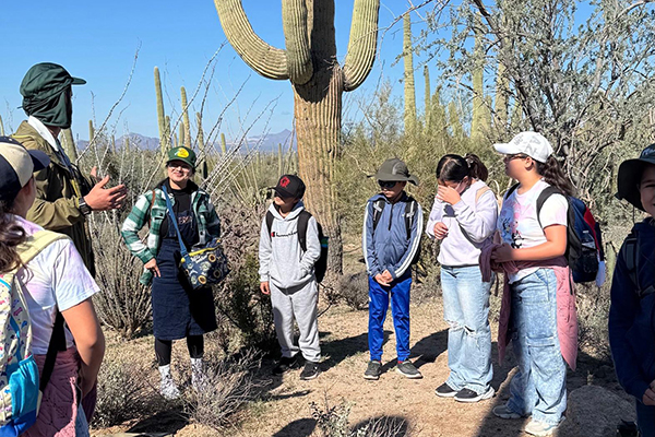 A teacher talks to his students as they stand around on a desert trail