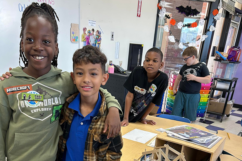 Two boys pose together in front of the bridge they made across their desks