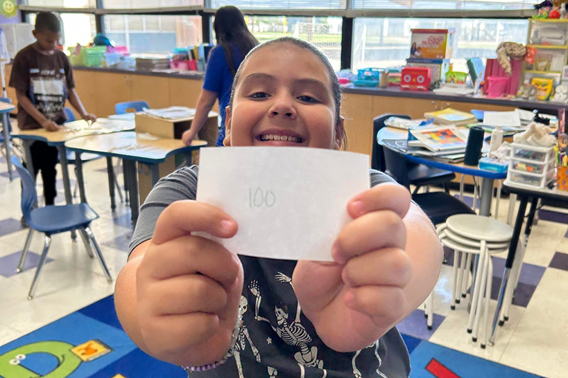 A girl smiles as she holds a piece of paper with 100 written on it up to the camera