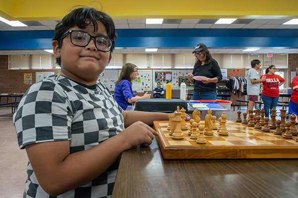 A boy wearing glasses and a checked shirt smiles as he plays chess