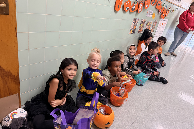 A group of young students sit on the floor in the hallway with their Halloween candy buckets