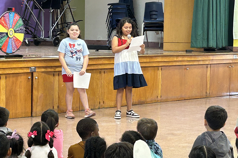 Two girls stand in front of the stage reading from pieces of paper